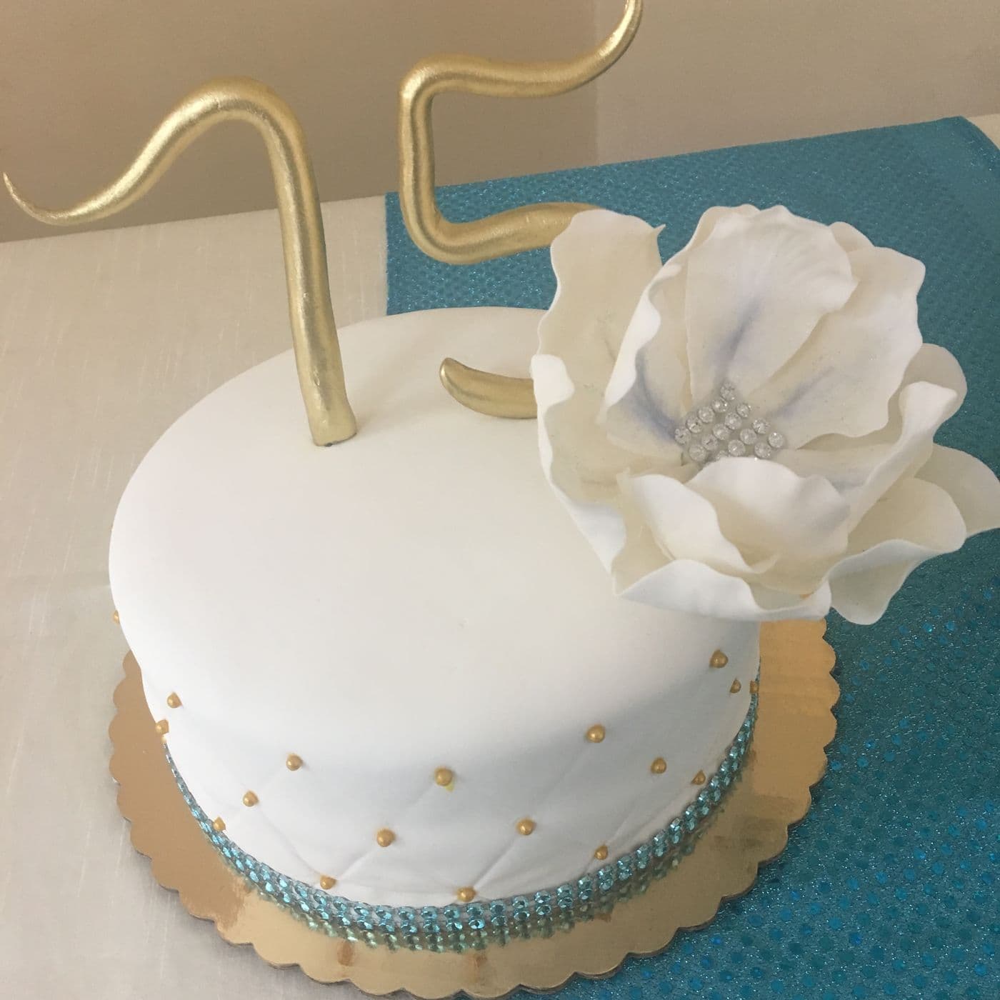 A 75th birthday cake in white fondant with a gold hand-made number topper, a large white sugar peony and teal crystal banding