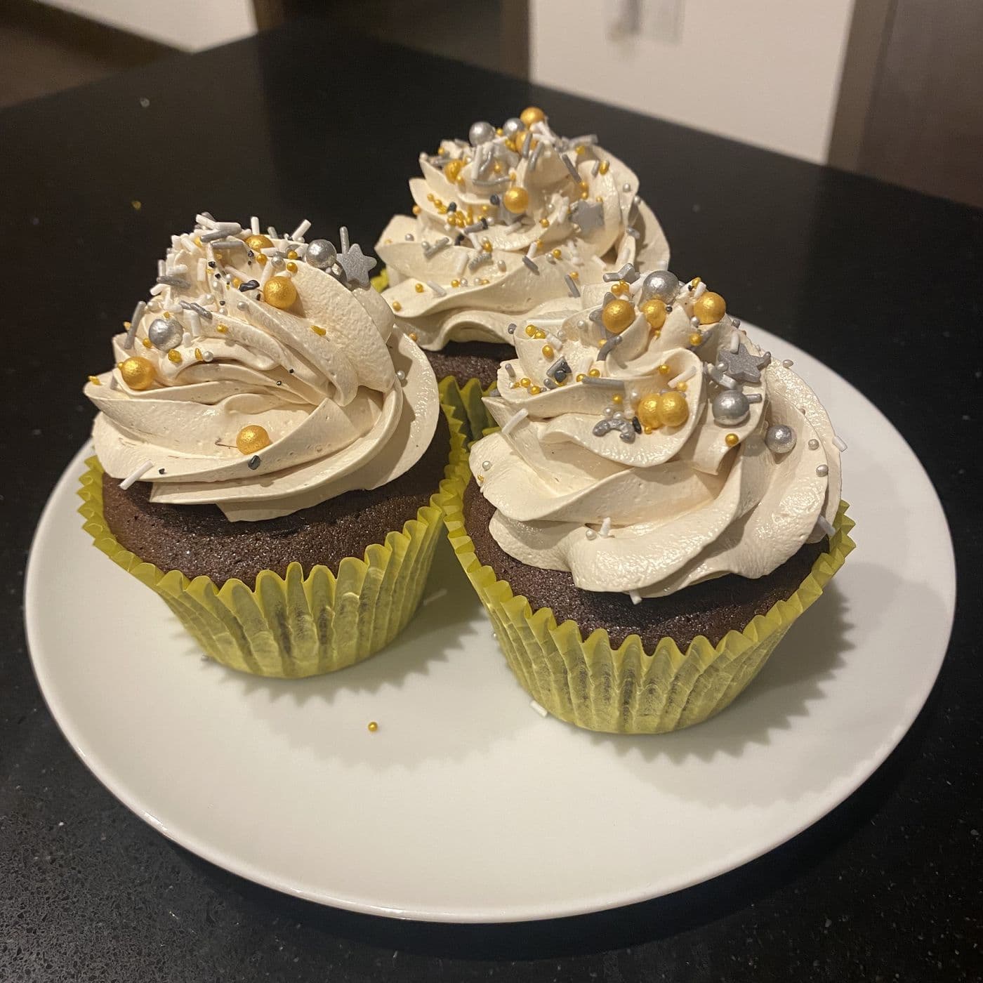 Three chocolate cupcakes in yellow cases on a white plate, topped with tall piped buttercream swirls and gold and silver sprinkles