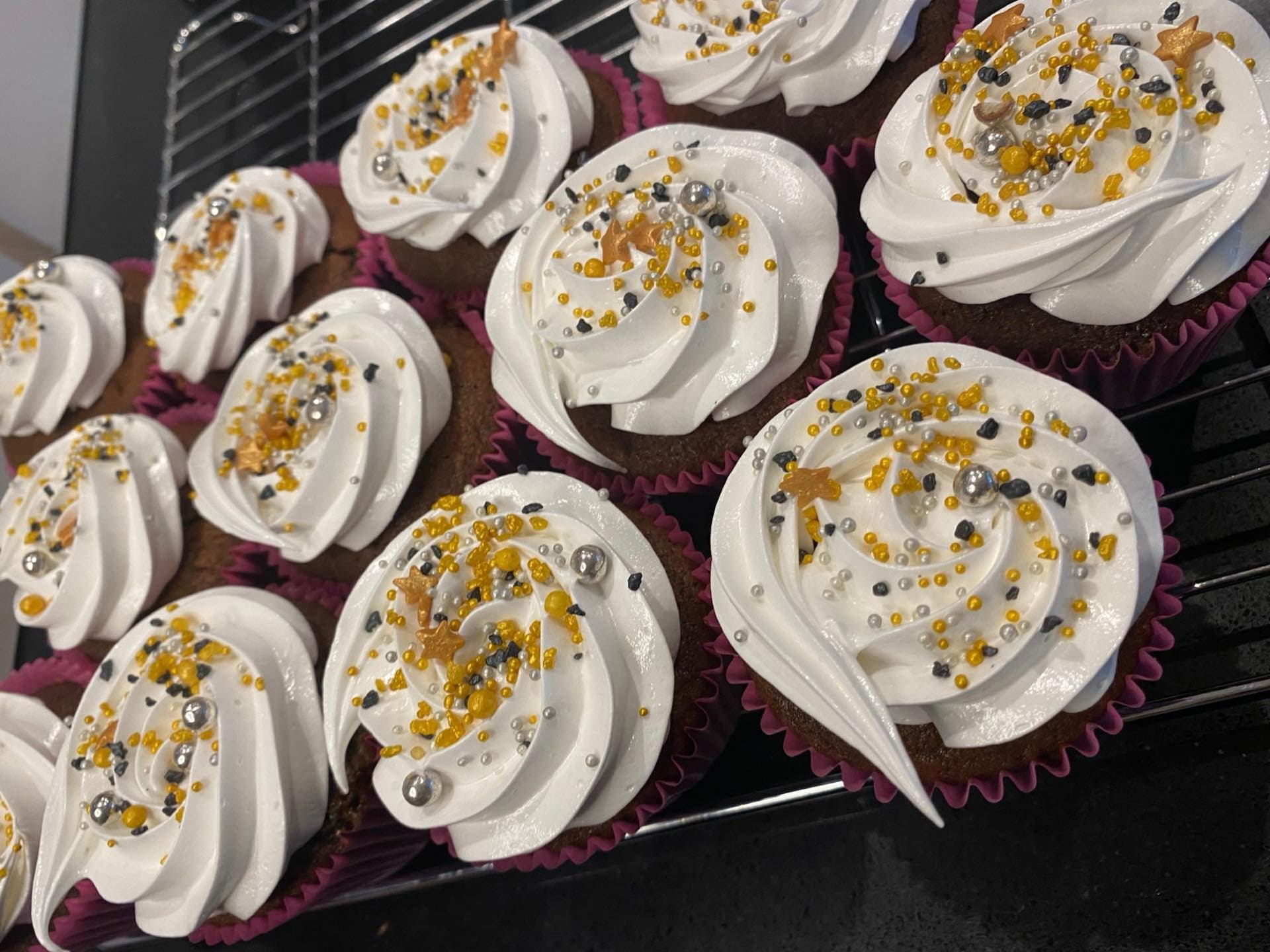 A full batch of hand-piped cupcakes with white buttercream swirls and gold, silver and black pearls, cooling on the rack