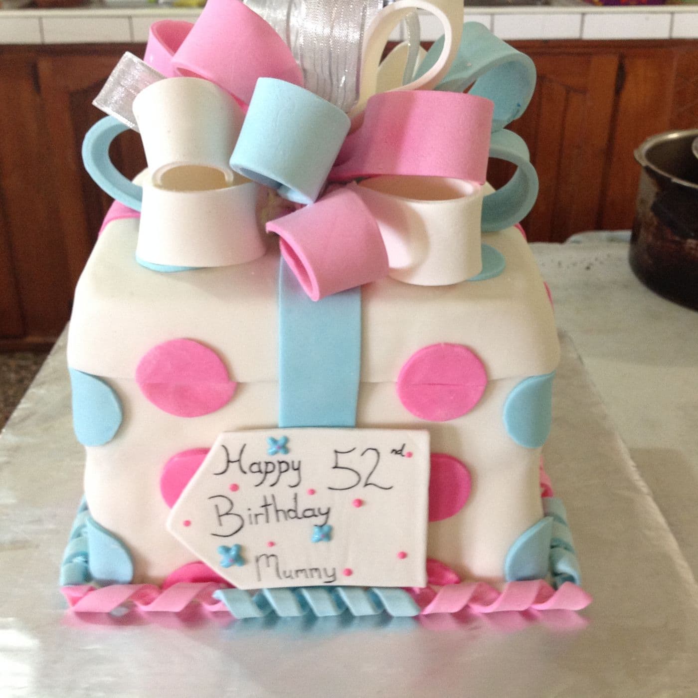 Square birthday cake decorated as a wrapped present with a pink and blue fondant loop bow, polka dots and a hand-written plaque
