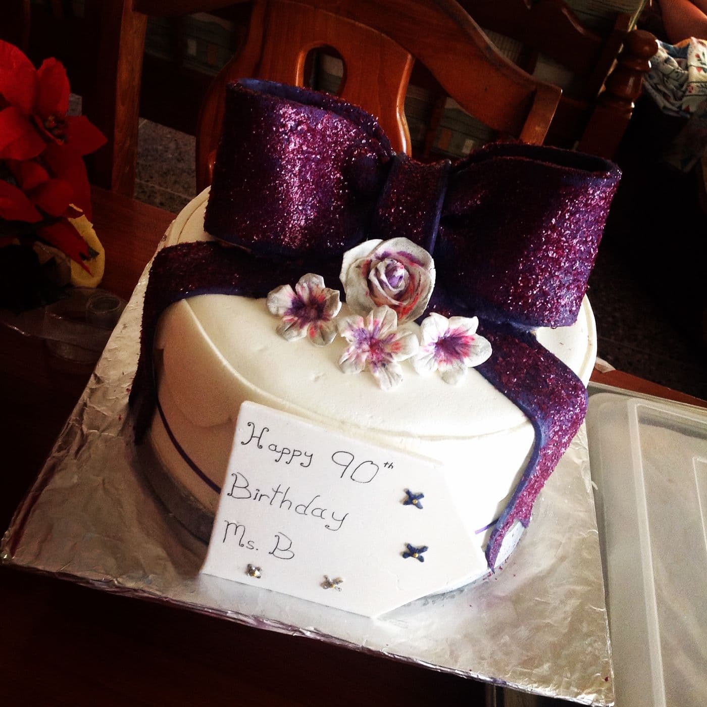 90th birthday cake in white icing with a large purple glitter fondant bow, a hand-made sugar rose and blossoms brushed with purple, and a hand-written plaque