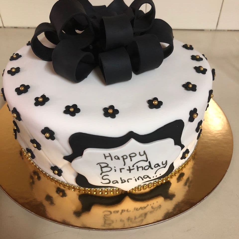 Birthday cake in white fondant with a black fondant loop bow, small black flowers with gold centres and a hand-piped name plaque, made to order in Peterborough