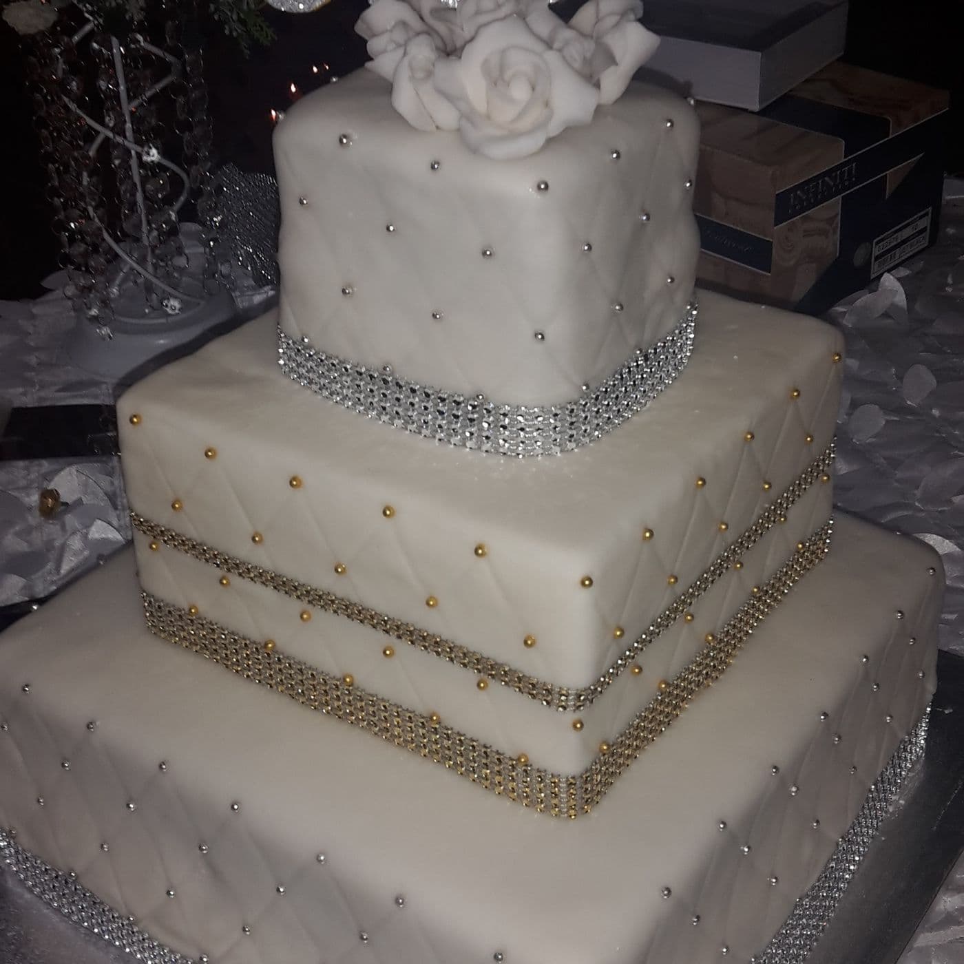 Close view of a three-tier square wedding cake showing quilted fondant, silver and gold crystal bands and dragée detailing on each tier